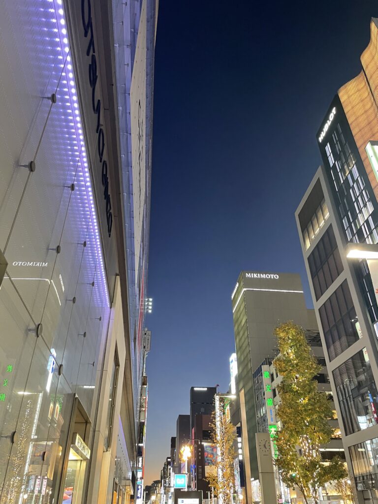 Ginza street at dusk with purple lights glowing on a building and city lights below a deep blue sky