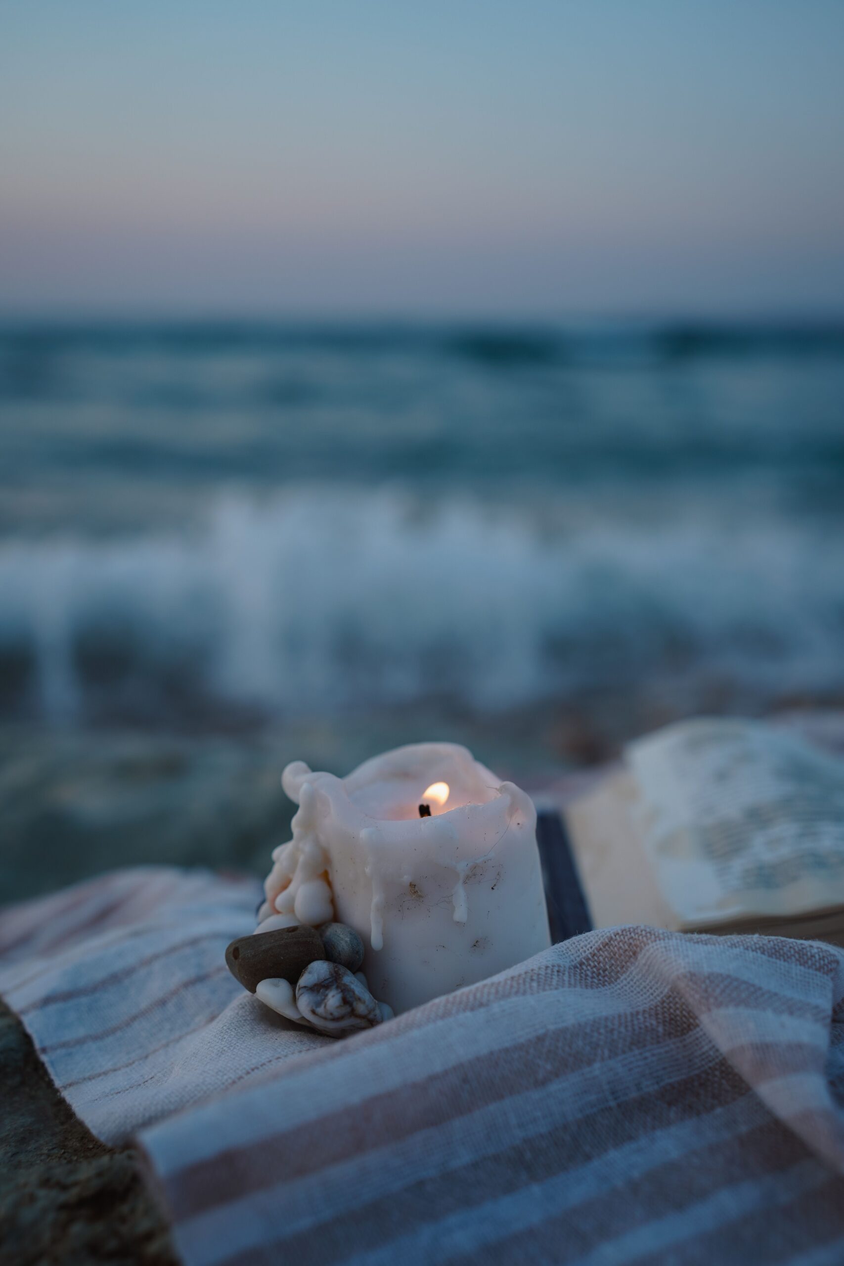 A single lit candle on a striped beach towel at twilight, with soft waves blurred in the background