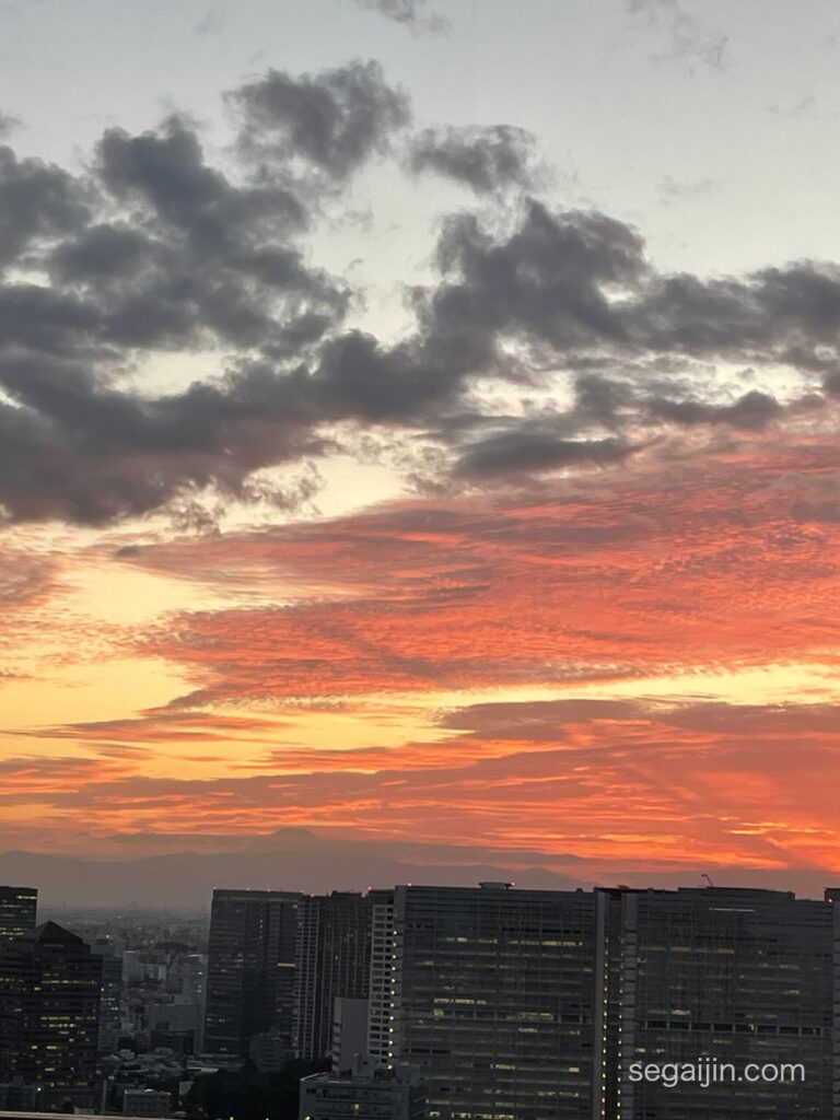 An orange and apricot-colored sunset sky over city buildings, with Mount Fuji faintly visible in the distance