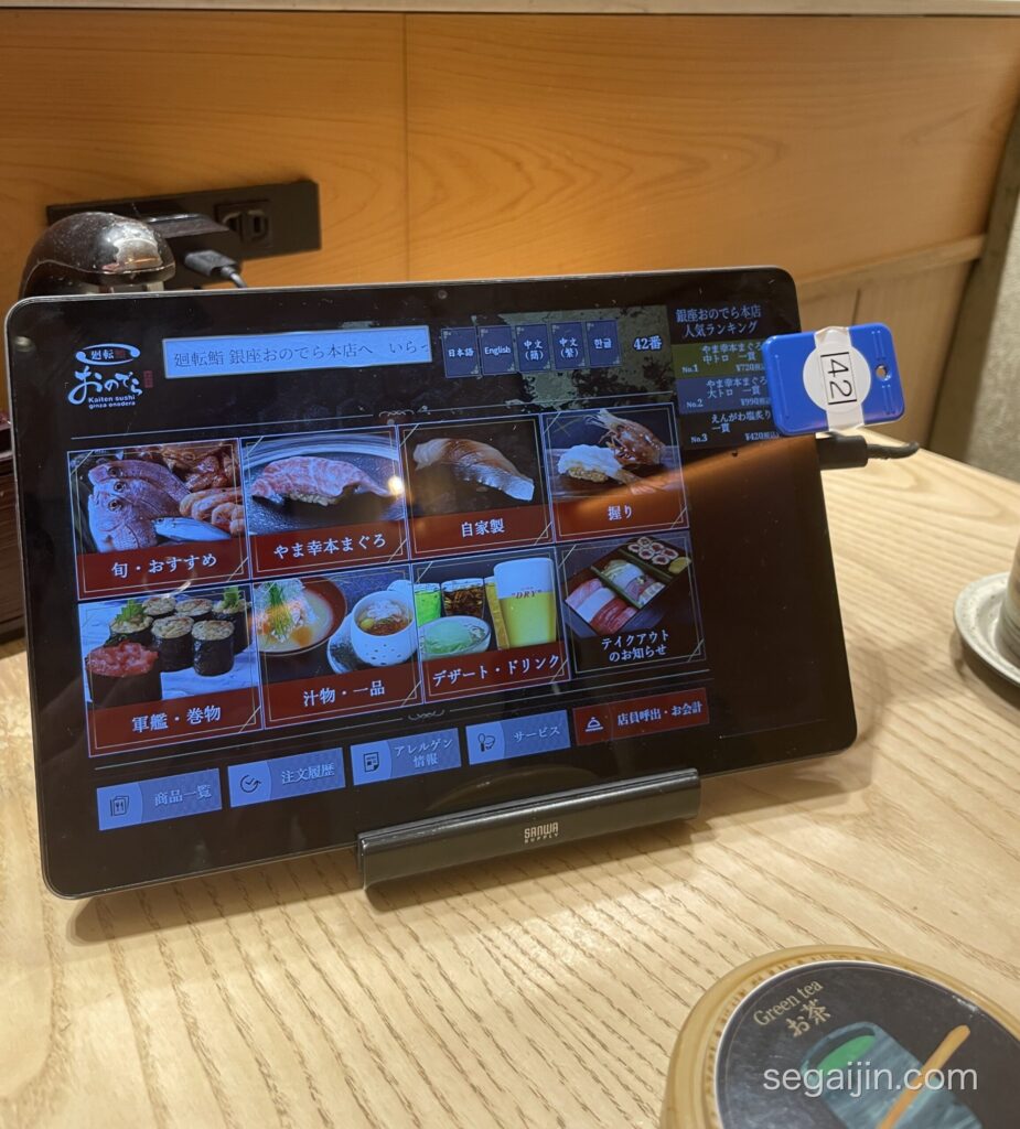 The ordering tablet at a conveyor-belt sushi restaurant, with language buttons and a numbered ticket tag